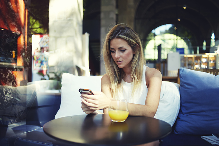 Portrait of a cute blonde female read something on her smart phone while sitting in modern coffee shop, beautiful young hipster girl using cell telephone while enjoying her free time in expensive cafe.
